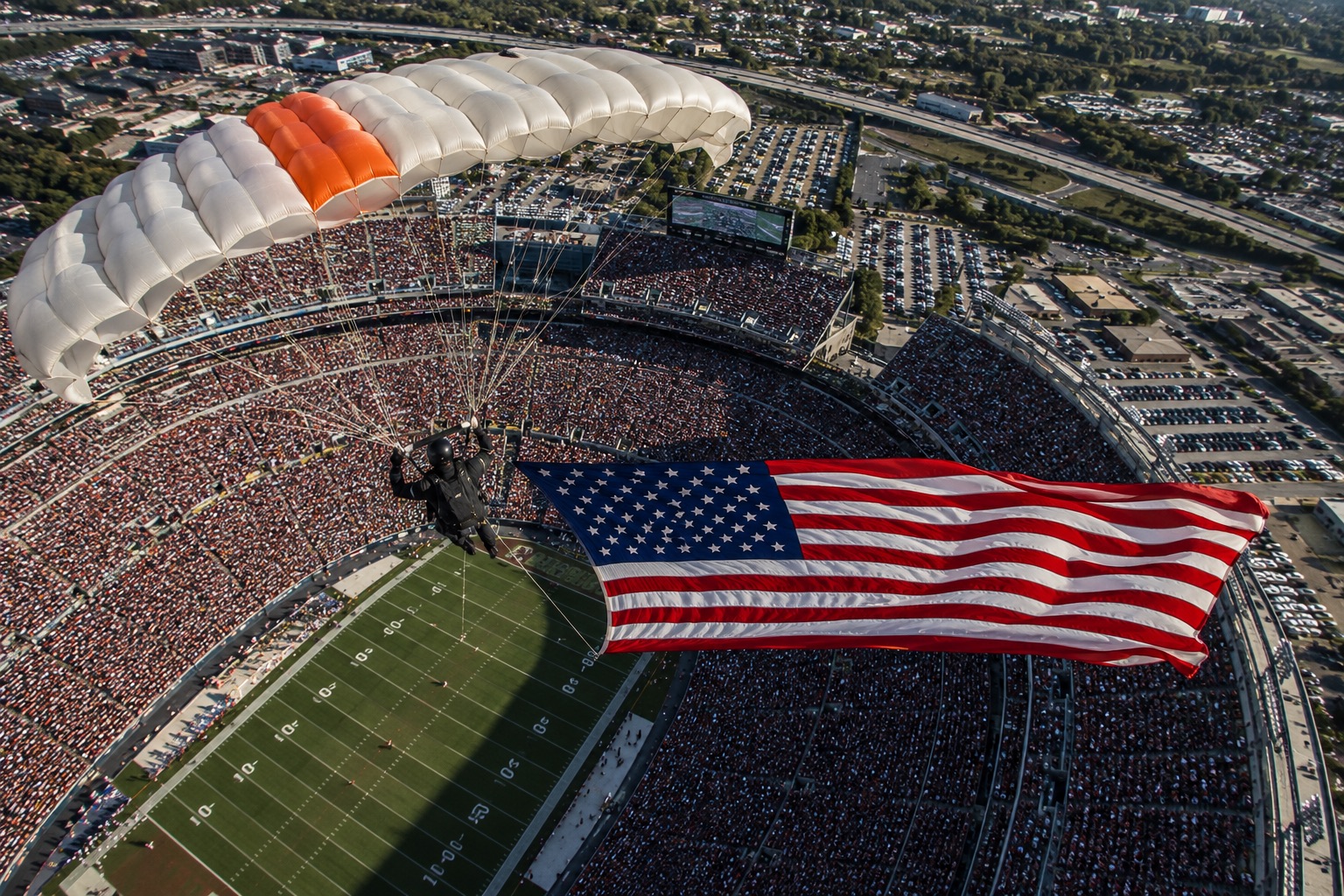 Parachutist trailing an American flag into a packed stadium bowl over the field